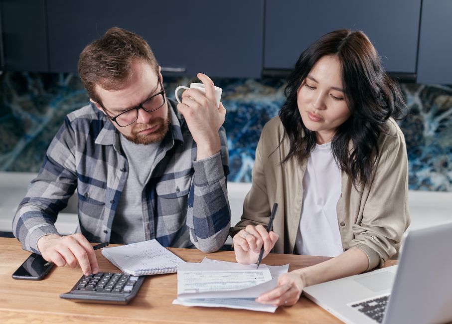Inflation personnelle : 3 calculs pour mesurer votre pouvoir d’achat A couple reviewing household bills and budget using a calculator and laptop at their kitchen table.