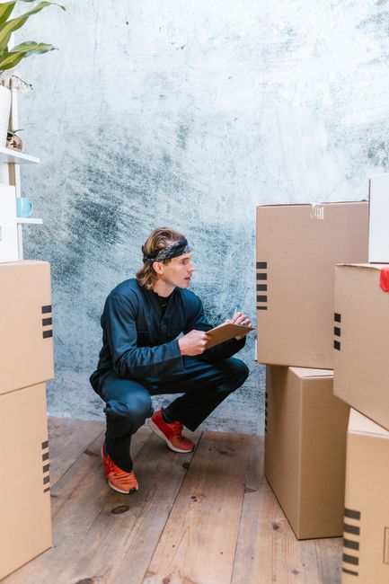 A man crouches to check shipping boxes, clipboard in hand, indoors.