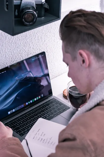 High angle of crop male student reading notes in notepad while using laptop and drinking coffee at desk