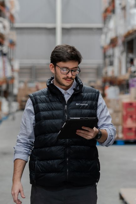 Young male in warehouse checking inventory on a tablet, surrounded by stacked boxes.