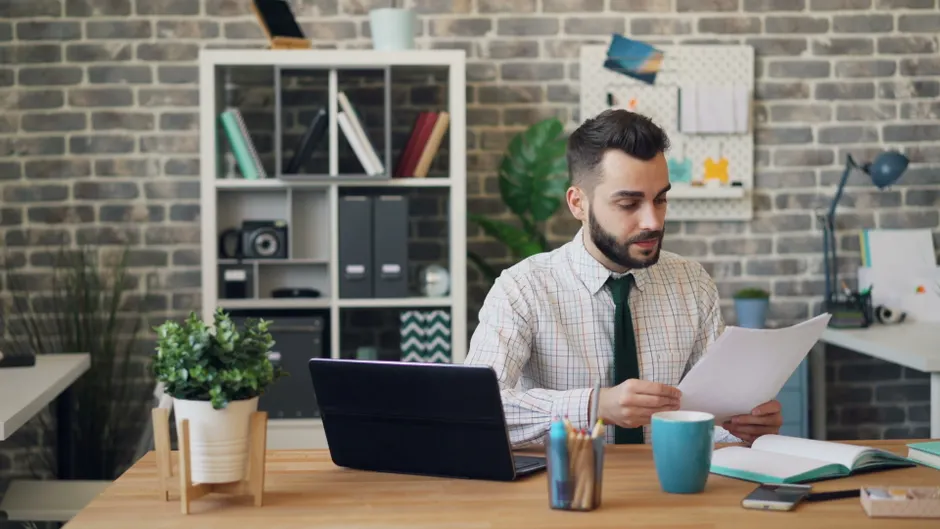 Jours de carence arrêt maladie : 3 jours dans le privé, 1 pour les fonctionnaires Young man reading papers at desk with laptop and office decor, focused on work.